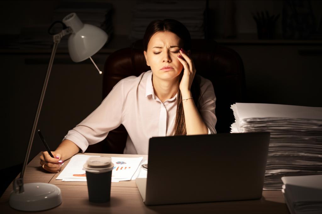A tired businesswoman sitting at her desk late at night in a dimly lit office, surrounded by papers and a laptop.